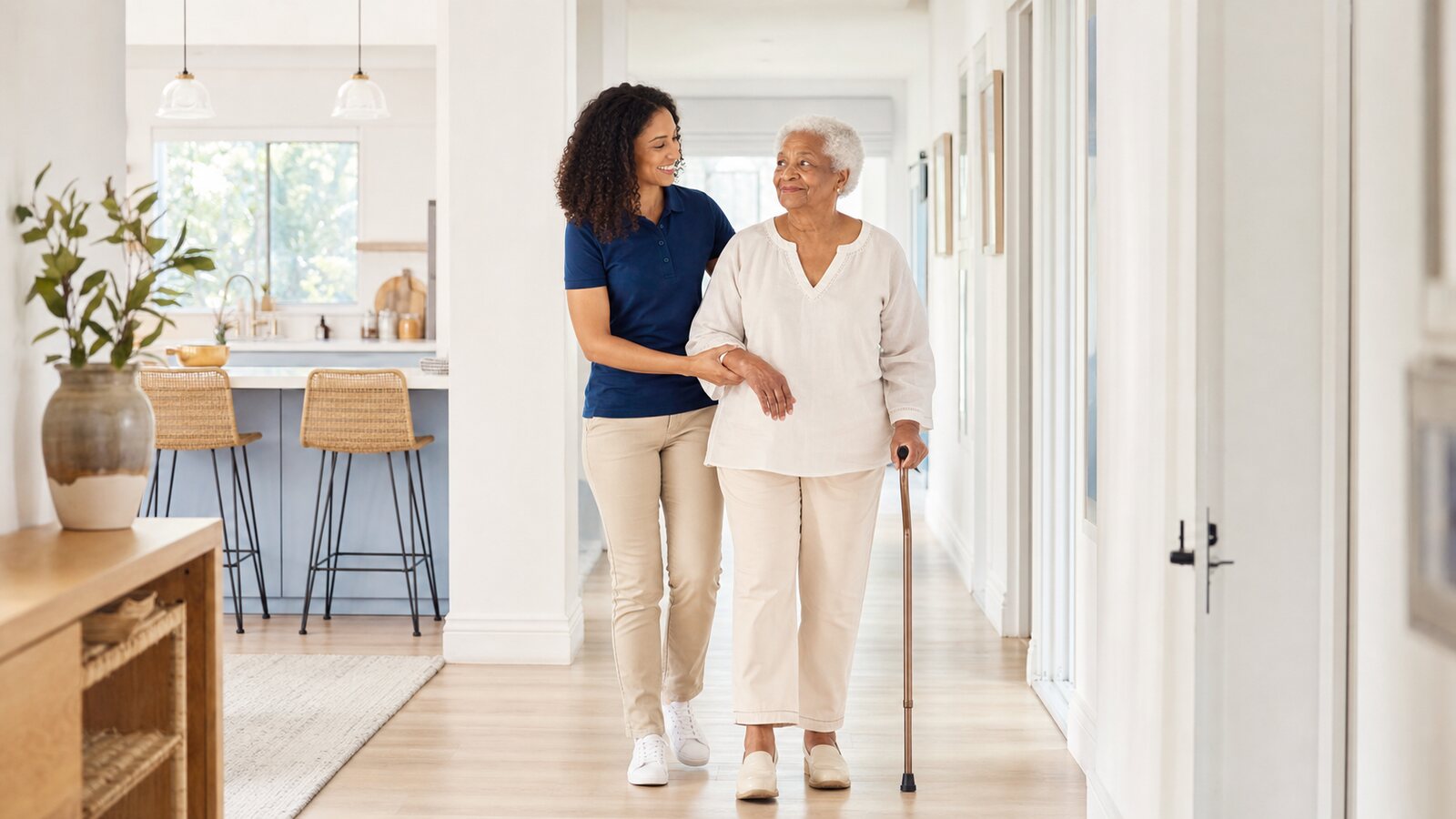 A caregiver supporting an older adult walking through a bright home